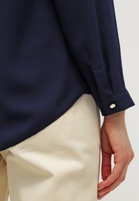 Námořnická modrá blůza s hladkým povrchem, s manžetou na knoflíky a šikmým lemováním. Kombinováno se světlými, volnočasovými kalhotami.