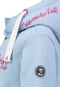 Lichtblau hoodie met witte trekkoorden, roze geborduurde tekst "Moin von der Küste," en een ronde patch met de tekst "Strand, Meer, Sonne Moin."