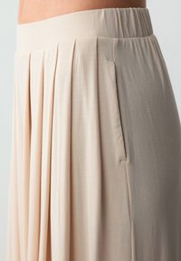 Lichtbeige geplooide rok met een zachte tailleband. De stof lijkt glad en lichtjes rekbaar, met parallelle plooien aan de zijkant.