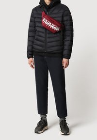 Sort pufferjakke med lynlås foran, parret med mørke jeans og sorte sko. Rød crossbody taske med markant hvid skrift.