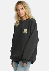 Felpa nera oversize con maniche lunghe, scollo a costine e una grafica beige del logo sul petto. Tessuto dalla consistenza delicata.