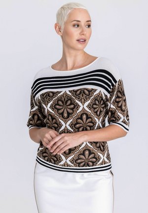 Femme aux cheveux blonds courts portant un haut à motifs floraux marron et noir avec des rayures noires et blanches, posant les mains jointes.