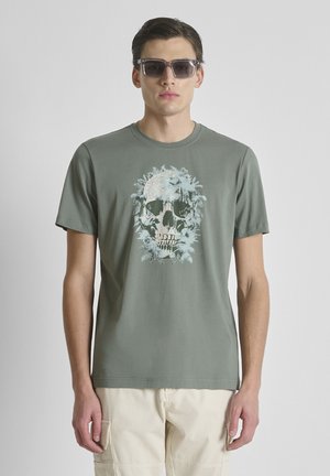 Hombre con camiseta gris verdosa con un gráfico de calavera y follaje, pantalones cortos cargo beige y gafas de sol oscuras, de pie frente a un fondo liso.