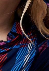 Barevná vzorovaná tkanina s diagonálními pruhy v modré, červené a bílé barvě. Obsahuje límec s knoflíky, který ukazuje dynamický design.