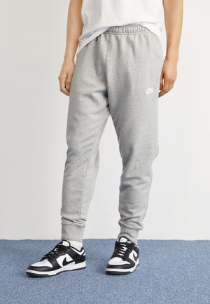 Mężczyzna ubrany w szare spodnie dresowe Nike i czarno-białe sneakersy Nike stoi na niebieskim wykładzinowym podłożu.