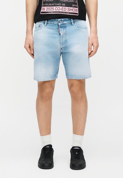 Homme portant un short en jean bleu clair, des chaussettes blanches, des baskets noires, et une partie d'un t-shirt noir à motif visible sur un fond uni.
