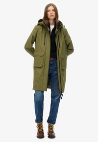 Parka impermeable de color verde oliva con capucha, dos grandes bolsillos frontales, cremalleras laterales y detalles en negro, combinado con jeans azules y botas marrones.