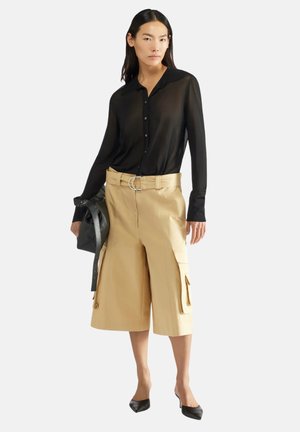 Sort gennemsigtig skjorte med knapper, beige cargo shorts med sidelommer og stort rundt bæltespænde, sorte spidse flade sko og en sort taske.