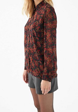 Vrouw die een zwarte blouse met knopen en rode bloemenprint draagt, grijze shorts en doorzichtige zwarte panty's, staand voor een witte achtergrond.