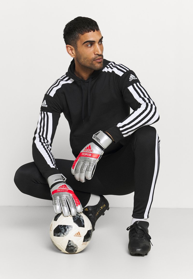 Haut à capuche noir avec des rayures blanches, leggings noirs, gants de gardien argentés et rouges, chaussures de foot noires et un ballon de soccer à motifs.