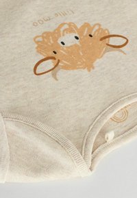 Ekologisk bomullsoveralls i ljus beige med en lustig brungrafik av ett djur och tryckknappar i nederkant. Mjuk textur, rund halsringning.
