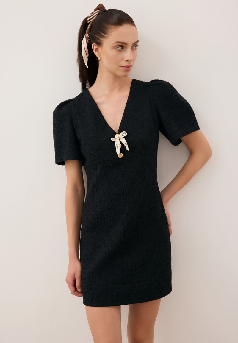 Zwarte korte jurk van een textuurstof, met V-hals, pofmouwen en een decoratieve strik met een knoopaccent aan de voorkant in het midden.