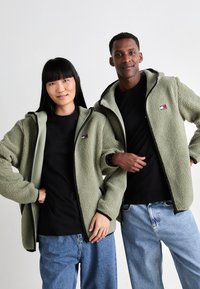 Twee mensen in groene fleecjacks, zwarte shirts en blauwe spijkerbroeken staan tegen een effen witte achtergrond.