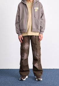 Gray zip-up hoodie with drawstrings, beige t-shirt underneath, and brown cargo pants with multiple pockets. Footwear: brown sneakers with white accents.