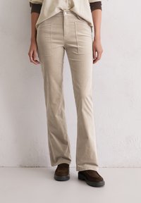 Beige corduroy broek met een uitlopende pasvorm, voorzien van patchzakken aan de voorkant en een subtiele gestructureerde oppervlakte, gedragen met bruine suède schoenen.