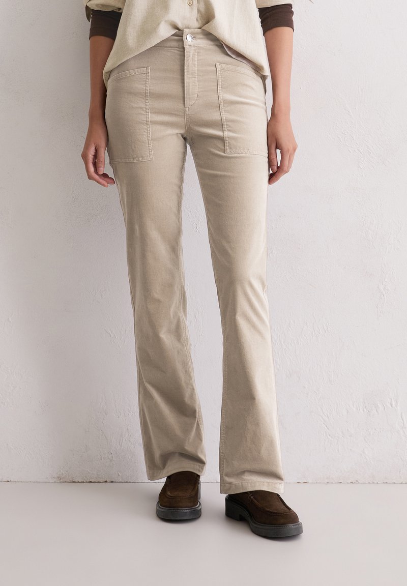 Beige corduroy broek met een uitlopende pasvorm, voorzien van patchzakken aan de voorkant en een subtiele gestructureerde oppervlakte, gedragen met bruine suède schoenen.