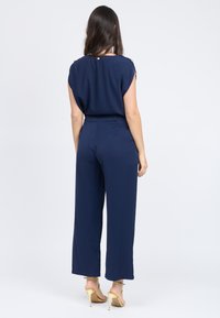 Navy blauwe jumpsuit met een ontspannen pasvorm, wijde broekspijpen en decoratieve gouden knopen op de schouder. Glad materiaal en een op maat gemaakte ontwerp.