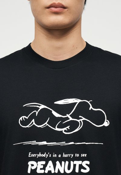 Άτομο που φοράει μαύρο πουκάμισο με λευκό περίγραμμα του Snoopy που τρέχει και το κείμενο «Όλοι βιάζονται να δουν τα PEANUTS.»