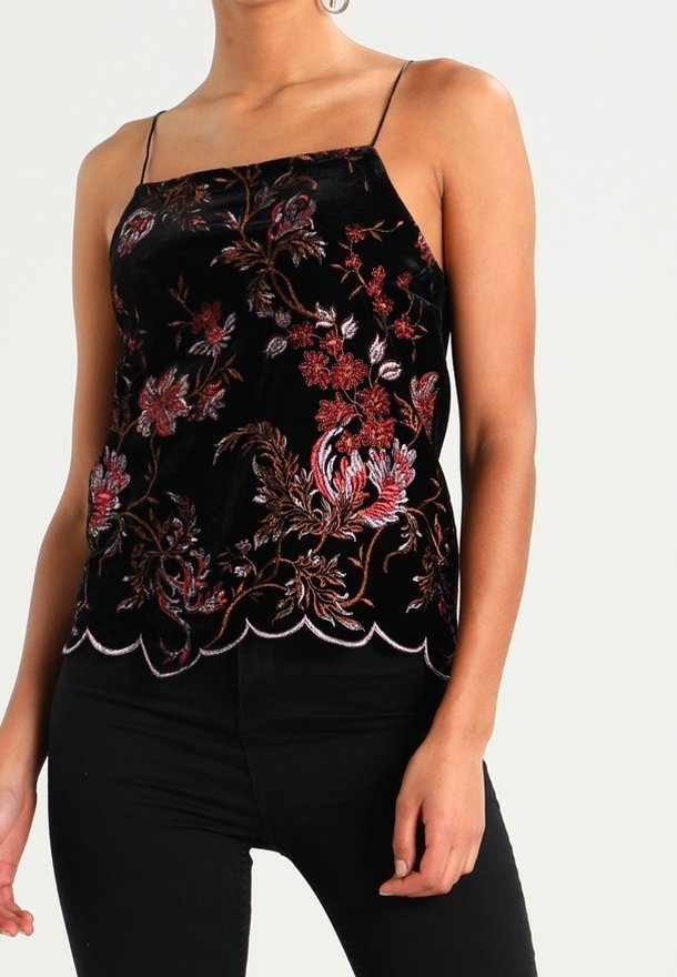 Femme portant un caraco en velours noir avec broderies florales rouges et roses et fines bretelles, associé à un pantalon noir.
