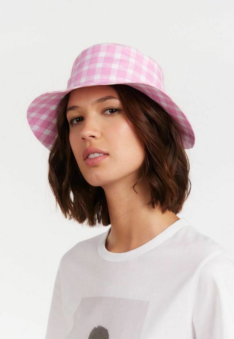Chapeau seau en vichy rose et blanc avec un large rebord et un tissu doux. Porté avec une chemise blanche basique, créant un look décontracté.