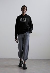 Crna kapuljača s metalnim logotipom, sivi elastični hlače s ankle, i crni tenisice. Model stoji protiv neutralne pozadine.