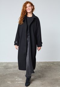 Sort uldfrakke med en struktureret silhuet, stor krave og knaplukning ved manchetten. Båret over en sort top og grå jeans med opsmøg.
