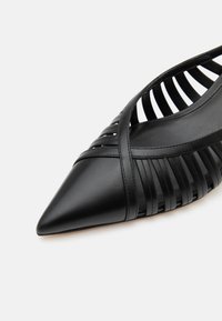 Chaussure plate noire à bout pointu avec des brides croisées et des côtés ouverts présentant des fentes verticales, en cuir lisse.