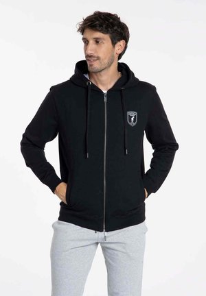 Sweat-shirt à zip noir avec une poche frontale, capuche à cordon et logo brodé. Tissu doux avec des manches et un ourlet côtelés. Associé à un pantalon de jogging gris.