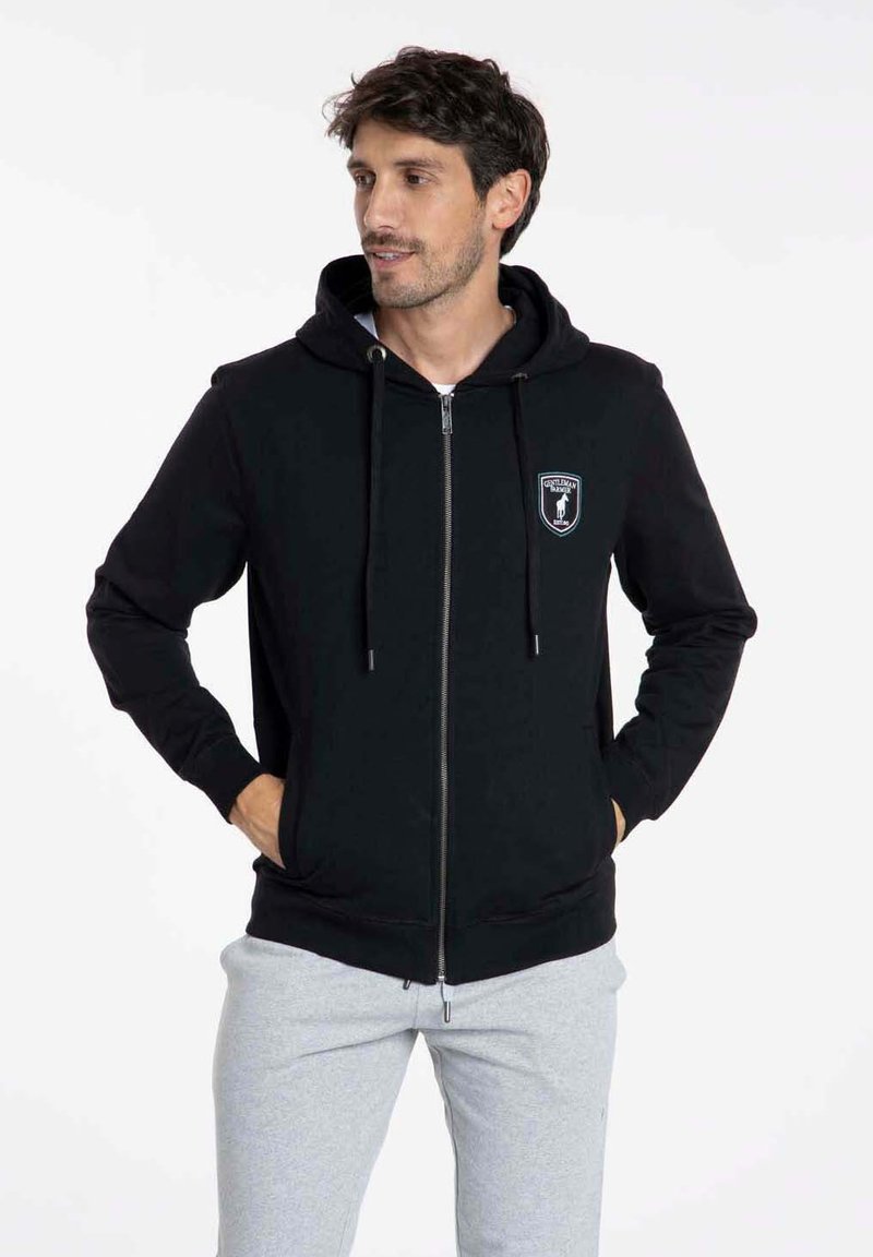 Sweat-shirt à zip noir avec une poche frontale, capuche à cordon et logo brodé. Tissu doux avec des manches et un ourlet côtelés. Associé à un pantalon de jogging gris.