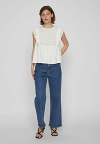 Top peplum blanco con patrón de ojal y mangas abullonadas, combinado con jeans azules de campana. Sandalias negras de tiras completan el atuendo.