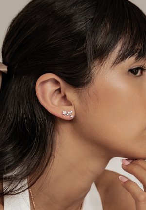 Frau mit dunklen Haaren, die kleine silberne sternförmige Ohrringe trägt und ihr Kinn auf die Hand mit gepflegten Nägeln stützt.