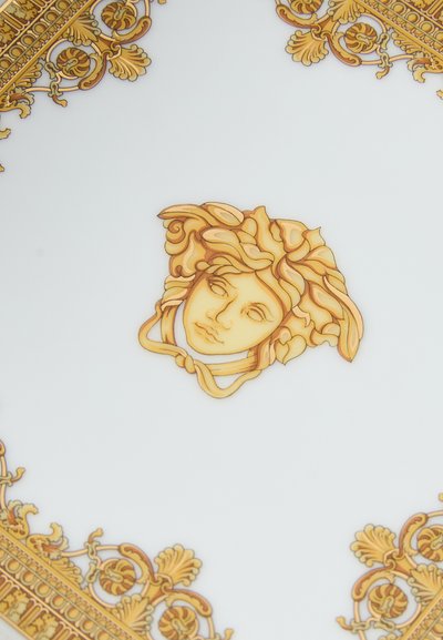 Apaļā porcelāna šķīvis ar centrālu zelta un oranžu dizainu, attēlojot seju ar izsmalcinātiem matiem, ko ieskauj sarežģītas zelta raksti malas daļā.