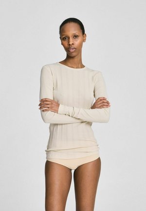 Ženska z rokami prekrižanimi na prsih, oblečena v bež rebrast dolg rokav in ustrezno spodnje perilo, stoji pred enobarvnim svetlo sivim ozadjem.