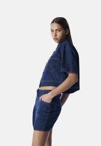 Veste en denim coupée avec des manches courtes et deux poches avant, associée à un short en denim taille haute, le tout dans un lavage bleu foncé.