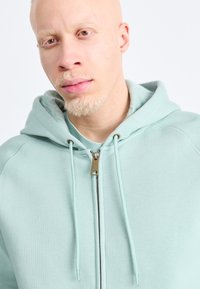 Svetlomodrá mikina na zips s viazaním, s hladkým povrchom a strieborným zipsom. Zmiešaná tkanina s voľným strihom.