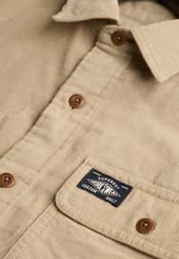 Chemise beige en velours côtelé avec des boutons marron et une étiquette rectangulaire bleu marine sur la poche poitrine indiquant "Superdry Workwear Custom Built."