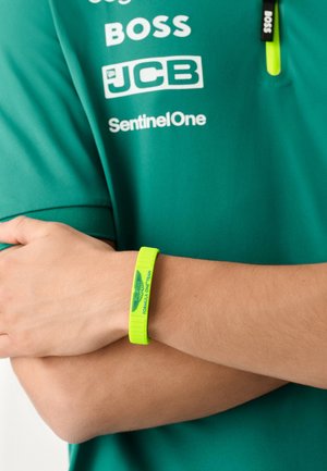 Grünes Polohemd mit weißen Logos und einem leuchtend neongelben Silikonarmband mit der Aufschrift "FORMULA ONE TEAM" und Flügel-Logo.