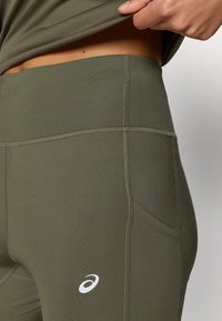 Λεγίνες σε ελιά πράσινο χρώμα από ελαστικό υλικό, με λεία υφή, πλευρική τσέπη και αντίθετο λευκό λογότυπο στο ισχίο.