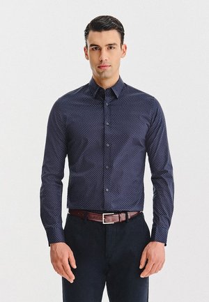 Mężczyzna w dopasowanej granatowej koszuli w małe białe kropki, czarnych spodniach i brązowym pasku, stojący frontalnie z neutralnym wyrazem twarzy.