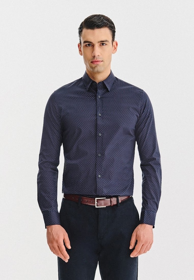 Mężczyzna w dopasowanej granatowej koszuli w małe białe kropki, czarnych spodniach i brązowym pasku, stojący frontalnie z neutralnym wyrazem twarzy.