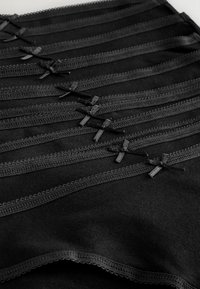 Czarna tkanina z kilkoma poziomymi pasami, ozdobiona delikatnym koronkowym wykończeniem i małymi materiałowymi kokardkami, tworząc teksturowany, warstwowy efekt.