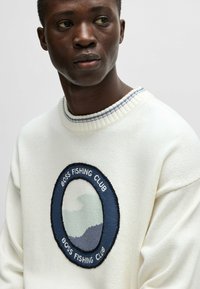 Maglione bianco con colletto a coste, dotato di una toppa ricamata circolare. La toppa presenta sfumature di blu e verde con il testo "BOSS FISHING CLUB".