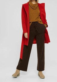 Manteau rouge en laine, haut marron en tricot, pantalon marron à jambes larges, et bottines beiges à la cheville. Le manteau présente une capuche et un design ouvert à l'avant.