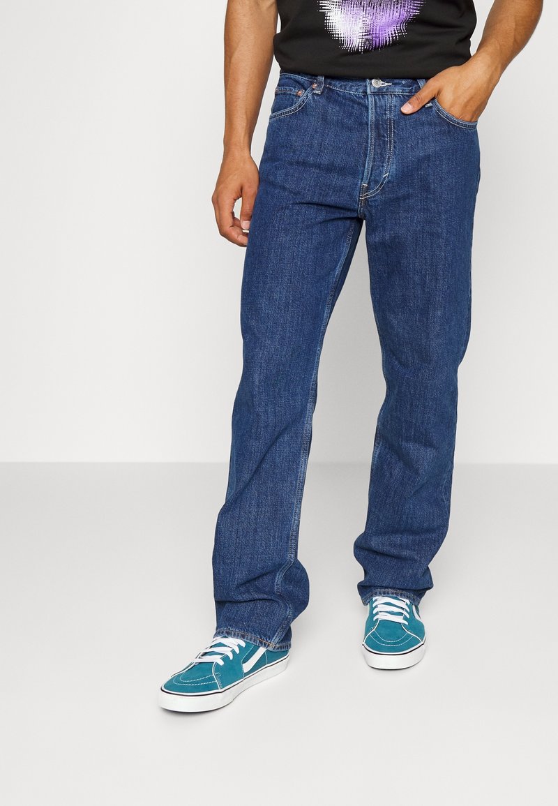 Homme portant un jean bleu, un t-shirt graphique noir et violet, et des baskets bleu sarcelle, debout avec une main dans la poche.