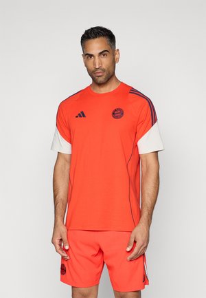 Φωτεινό πορτοκαλί αθλητικό μπλουζάκι με λευκά επενδυμένα μανίκια, ναυτικές μπλε ρίγες και λογότυπο στο στήθος; σε συνδυασμό με αντίστοιχα πορτοκαλί σορτς.