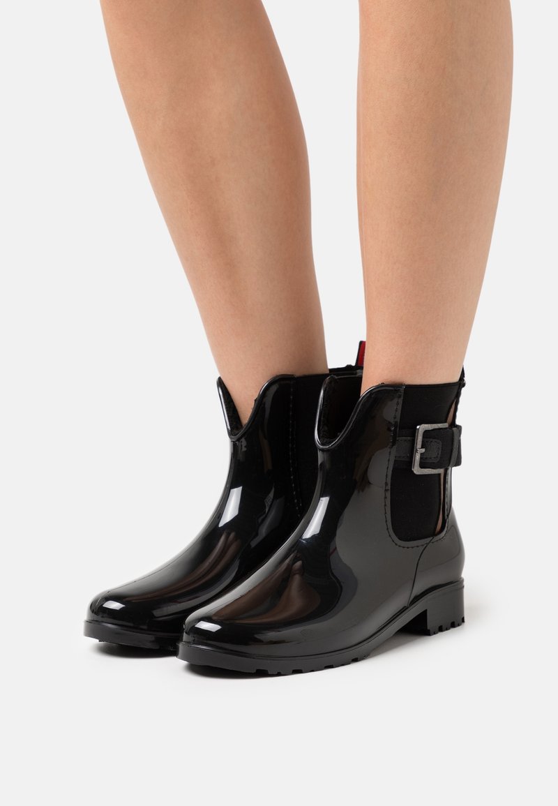 Bottes de pluie noires brillantes à la cheville avec boucle latérale portées sur des jambes nues, montrant un talon léger et un design à bout arrondi.