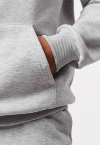 Siva dukserica s kapuljačom, džepom u obliku klokana, rebrastim manžetnama i rubom. Mekana tekstura i neupadljiva oznaka marke sa strane.