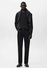 Črn pulover, obložen z sivim brezrokavnikom iz pletenine, v kombinaciji s črnimi ozkimi hlačami. Model nosi črne čevlje in stoji pred belim ozadjem.