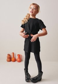 Sort tekstureret kort jumpsuit med knyttet talje, parret med sorte leggings og ankelstøvler. Farverige keramiske vaser i baggrunden.
