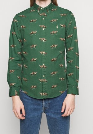 Zielona koszula na guziki z powtarzającym się nadrukiem koni i dżokejów, noszona z niebieskimi dżinsami, na osobie stojącej na jednolitym tle.